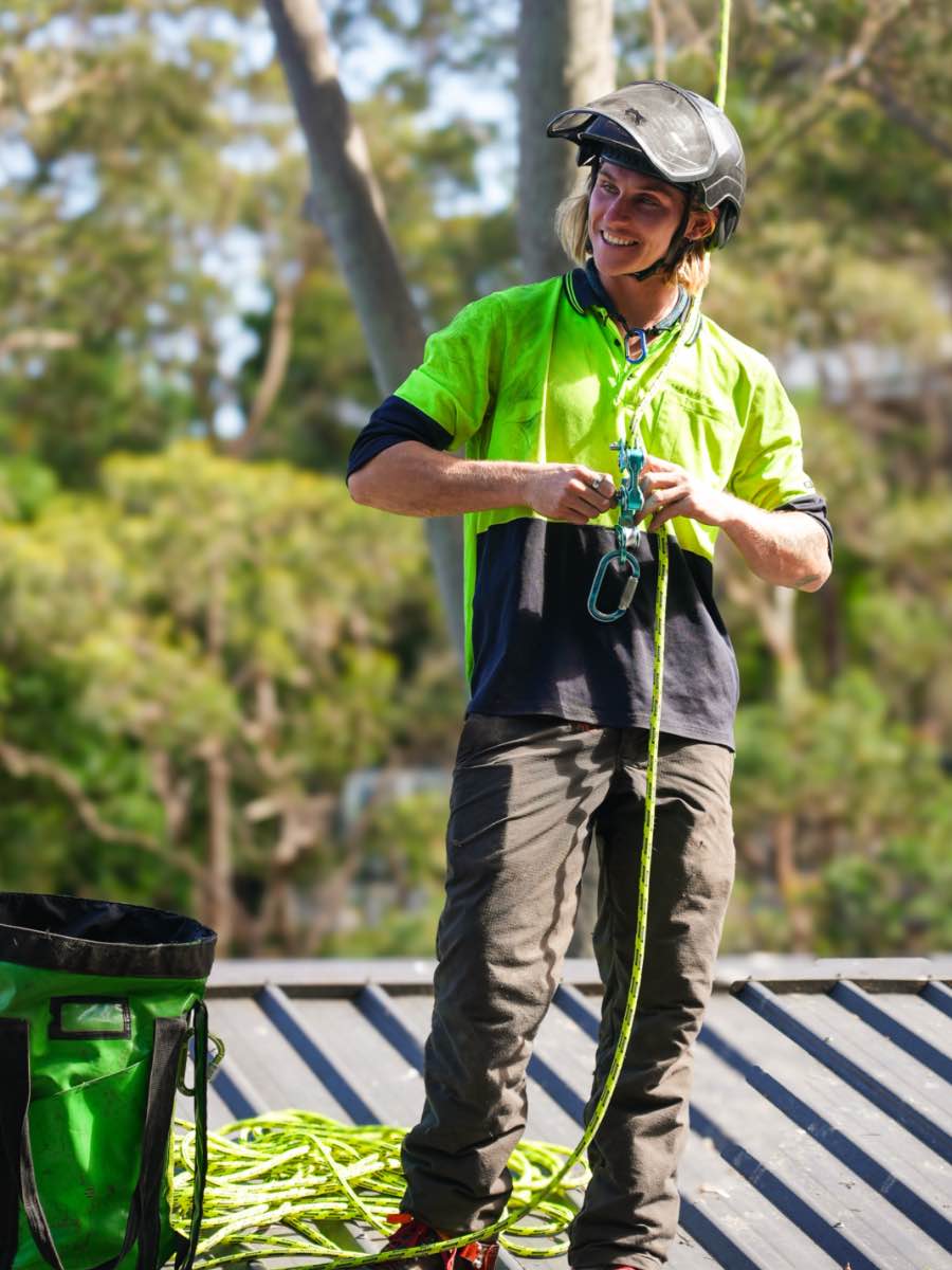 Smiling climber in a helmet and hi-vis clipping into a rope on a metal roof, with a rope bag and coiled line at his feet
